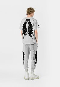 Camiseta gris con gráfico de alas negras en la parte posterior, combinada con pantalones deportivos a juego que presentan un diseño de alas similar y un detalle de correa lateral. Botas blancas.