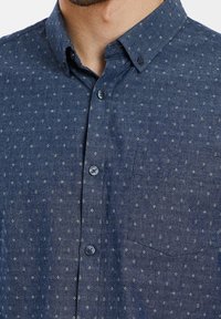 Gros plan d'un homme portant une chemise bleue à boutons avec de petits motifs floraux blancs et une poche poitrine.