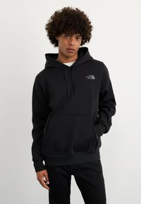 Felpa nera con cappuccio realizzata in tessuto morbido, con tasca a marsupio e un piccolo logo grigio. Il modello la indossa con jeans neri su uno sfondo neutro.