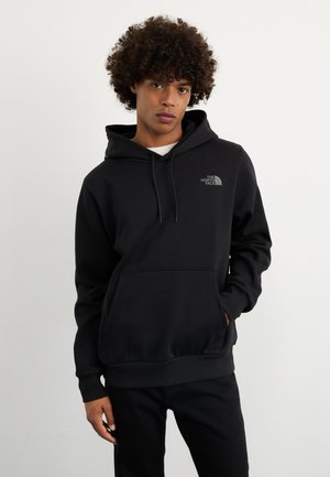 Felpa nera con cappuccio realizzata in tessuto morbido, con tasca a marsupio e un piccolo logo grigio. Il modello la indossa con jeans neri su uno sfondo neutro.