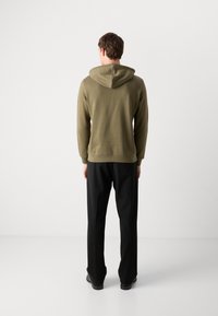 Olijfgroene hoodie met een achternaad, geribbelde manchetten en een capuchon met koord, gecombineerd met zwarte broeken en zwarte schoenen. Glad stoffenoppervlak.