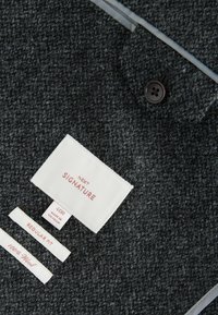Ciemnoszary, teksturowany materiał wełniany z guzikami i kilkoma metkami: "NEXT SIGNATURE," rozmiar "40R," "REGULAR FIT," oraz "100% Wełna."