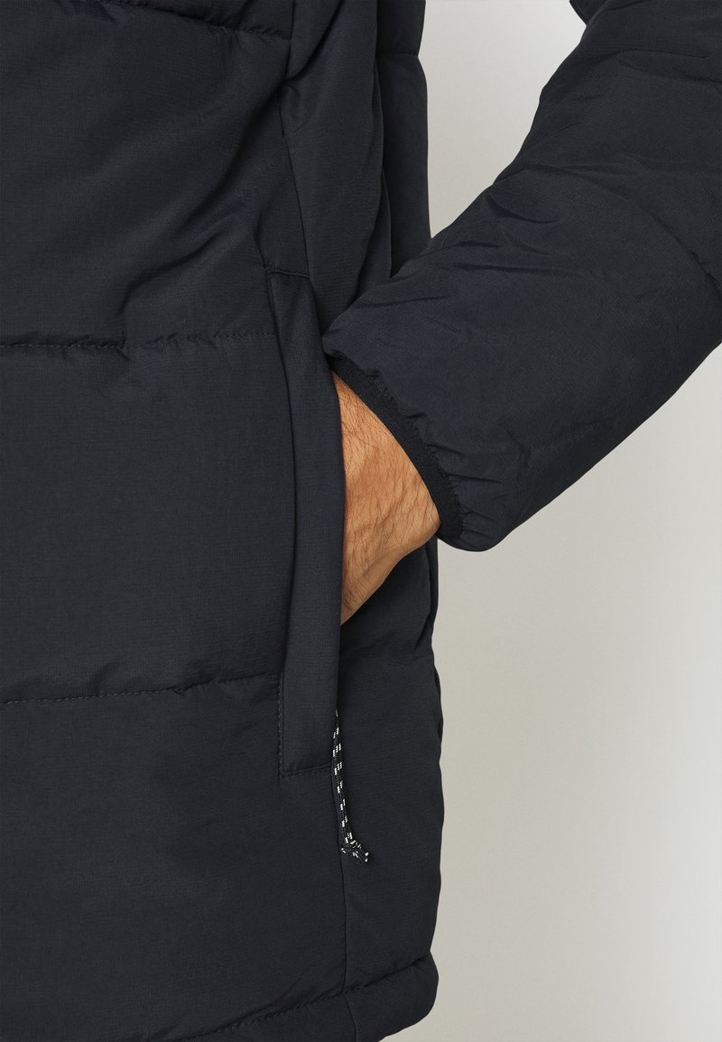 Giacca puffer blu navy con design trapuntato, tessuto testurizzato e polsini elasticizzati; mano nella tasca con dettaglio del cordino visibile.