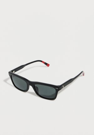 Schwarze, rechteckige Sonnenbrille mit dunklen Gläsern und roten Akzenten an den Bügelenden, die auf einer weißen Oberfläche liegt.