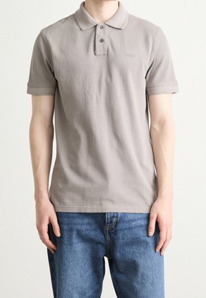 Mand iført en ensfarvet taupe kortærmet poloshirt med to knapper og blå jeans, stående mod en ensfarvet baggrund.