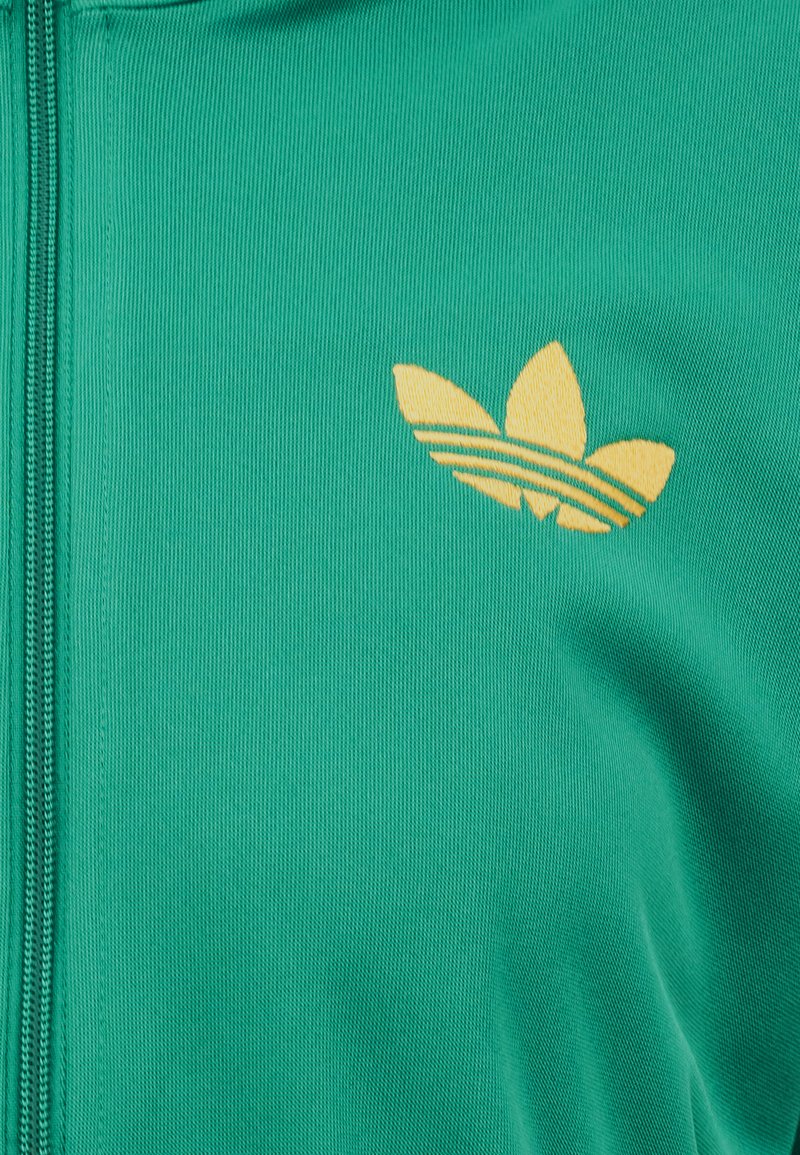 Grüner Stoff mit gelbem, gesticktem Adidas Trefoil-Logo, neben einer sichtbaren Reißverschlussnaht auf der linken Seite.
