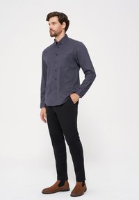 Chemise grise à manches longues avec un poignet à bouton et une poche poitrine, associée à un pantalon noir et des bottines brunes ; texture lisse et coupe ajustée.