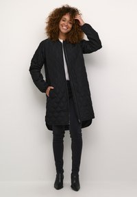 Manteau long noir matelassé avec fermeture éclair, présentant des motifs en losanges et des poches latérales, porté par-dessus un jean sombre et des bottines.