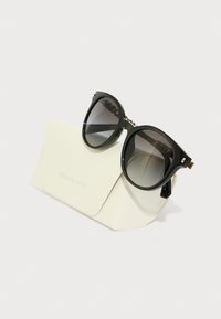 Okrúhle čierne slnečné okuliare s gradientovými sklíčkami, zlatými akcentmi, ležiace na bielom tekstúrovanom puzdre s embosovaným nápisom "MICHAEL KORS."
