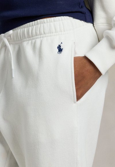 Κοντινό πλάνο ενός ατόμου που φοράει λευκή φόρμα με ρυθμιζόμενο κορδόνι και λογότυπο παίκτη πόλο σε ναυτικό μπλε, έχοντας το χέρι στην τσέπη.