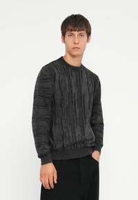 Black textured sweater with a ribbed neckline and cuffs, featuring vertical stripes and a subtle pattern, worn over dark pants.
