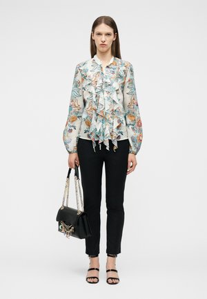 Woman wearing a floral ruffled blouse, black slim pants, black strappy heels, holding a black handbag with gold chain straps.