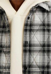 Rutig flanellskjorta i svart och vitt, med ett quiltat mönster och svarta tryckknappar, lager på lager över en krämfärgad hoodie.