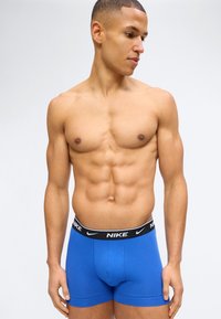 Αθλητικός άνδρας με κοντά μαλλιά που φοράει μπλε μποξεράκια Nike, στέκεται μπροστά σε απλό φωτεινό φόντο, κοιτάζοντας προς τα δεξιά του.