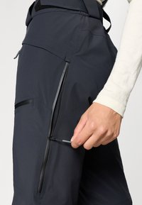 Zwarte waterdichte broek met zijzak met rits. Het materiaal lijkt glad en duurzaam. Een hand reikt naar de ritstrekkker. Subtiele accenten zichtbaar.