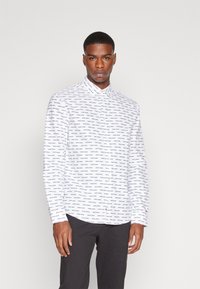 Camicia bianca con colletto, maniche lunghe e orlo curvo, decorata con un motivo di testo blu ripetuto. Indossata con pantaloni scuri.