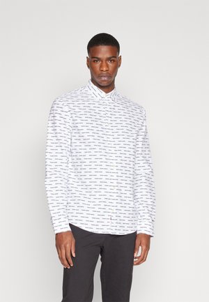 Camicia bianca con colletto, maniche lunghe e orlo curvo, decorata con un motivo di testo blu ripetuto. Indossata con pantaloni scuri.
