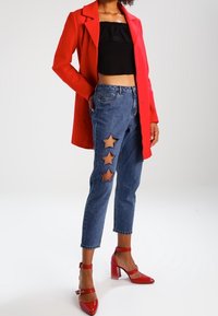 Rode lange jas, zwarte cropped top, blauwe jeans met steruitsparingen, en rode hoge hakken met enkelschoenen. Gladde texturen, casual ontwerp.