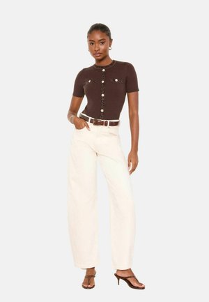 Vrouw staat met hand in zak, draagt een bruin, kort mouwig, knoopjesoverhemd, crèmekleurige, hoog geplaatste, wijde broekspijpen en bruine sandalen met hak.