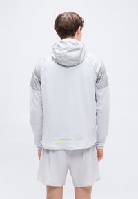 Ανοιχτό γκρι αδιάβροχο με κουκούλα, αντίθετες ασημί λεπτομέρειες στους ώμους και ανακλαστικό λογότυπο στο κάτω μέρος, κατασκευασμένο από συνθετικό υλικό.