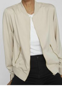 Beżowa kurtka bomberka z prążkowanym kołnierzem, złotym zamek błyskawicznym i bocznymi kieszeniami. Wykonana z gładkiej tkaniny, ma luźny krój i elastyczne mankiety.