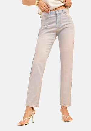 Kobieta w jasnych, prostych jeansach i beżowych sandałach na wysokim obcasie, trzymająca dół białego topu, stojąca na jednolitym tle.