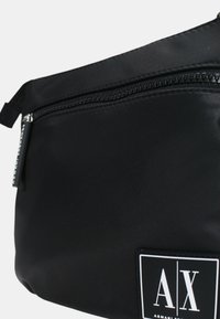 Borsa a tracolla nera in nylon con chiusura a zip, dotata di un evidente patch con logo bianco e texture liscia. Design compatto, leggera.