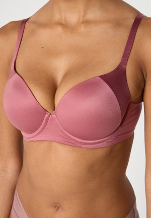 Kobieta nosząca dwukolorowy różowy usztywniany biustonosz z regulowanymi ramiączkami, pokazująca górną część tułowia i brzuch na jednolitym tle.