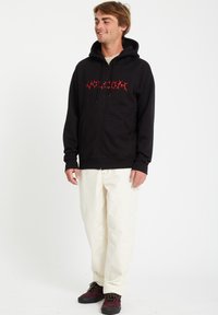 Zwarte rits hoodie met rode grafische tekst, voorzakken, gedragen met lichtbeige broek en bordeauxrode schoenen. Eenvoudige witte achtergrond.