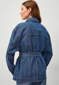 Giacca in denim di colore blu scuro con vita regolabile, colletto ampio e maniche lunghe. Presenta dettagli di cucitura e una vestibilità rilassata.