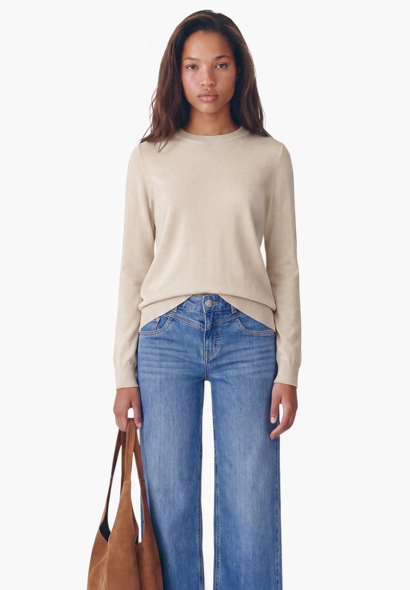 Jeune femme aux longs cheveux foncés portant un pull beige et un jean bleu, tenant un sac cabas en cuir suédé marron à ses côtés.