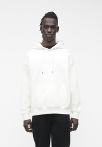 Sudadera con capucha blanca con bolsillo canguro, ajuste holgado, cordones y puños acanalados, combinada con pantalones oscuros.