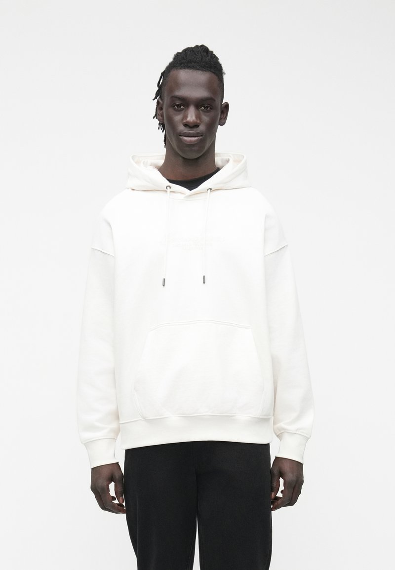 Sudadera con capucha blanca con bolsillo canguro, ajuste holgado, cordones y puños acanalados, combinada con pantalones oscuros.