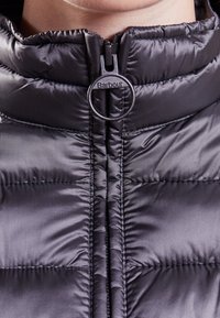 Schwarze Puffjacke mit hohem Kragen, ausgestattet mit einem Reißverschluss auf der Vorderseite und kreisförmigem Zubehör mit der Aufschrift "Barbour". Glatte, glänzende Textur mit gestepptem Muster.