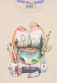Künstlerpalette mit einer gemalten Berglandschaft, Pinsel in einem Glas, eine Farbtube und getrocknete Blumen in einer Glasflasche auf einem beigen Stoff.