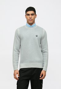 Ανοιχτό γκρι πλεκτό πουλόβερ με ραβδωτή υφή, στρογγυλή λαιμόκοψη και ένα μικρό λογότυπο στο στήθος, φορεμένο πάνω από ένα μπλε πουκάμισο με γιακά.