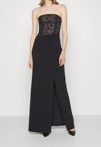 Banslös svart klänning med spetsöverdel och en draperad kjol med en sidoklyvning, accentuerad av en slät textur och en figursydd design.