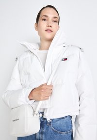 Giacca a vento bianca con zip, cappuccio e logo sul petto. Indossata sopra una camicia leggera. Tiene una piccola borsa a tracolla bianca con il marchio.