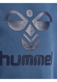 Niebieska bawełniana koszulka z dużym ciemnym logo o charakterystycznych kształtach i wzorach. Materiał ma teksturowaną powierzchnię i prążkowe wykończenie.