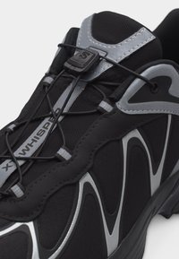 Μαύρο και γκρι αθλητικό παπούτσι με ανακλαστικές λεπτομέρειες, σύστημα γρήγορου δέσιμου, επενδεδυμένη γλώσσα και το κείμενο «X WHISPER» στο πάνω ύφασμα.