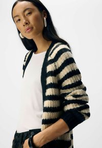 Schwarz-beige gestreifter Cardigan mit gestrickter Textur, offenem Frontbereich und gerippten Manschetten, getragen über einem weißen, figurbetonten Top.