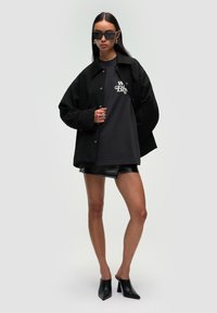 Chemise-chemise noire surdimensionnée superposée à un t-shirt graphique gris et une mini-jupe en simili cuir noire. Portée avec des mules à talons noirs et des lunettes de soleil.