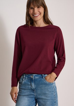 Femme souriante portant un haut bordeaux à manches longues et un jean bleu, une main dans la poche, devant un fond uni.
