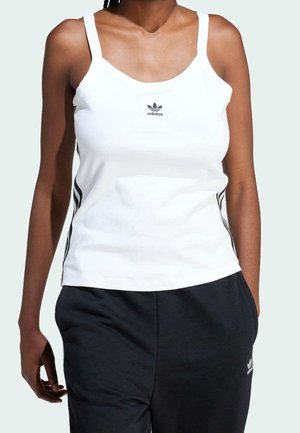 Kobieta w białym topie Adidas z logo i czarnych spodniach Adidas, stoi z jedną ręką w kieszeni na jasnym tle.