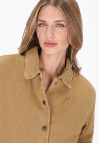 Olajzöld kabát klasszikus gallérral, az egyik oldalon horgony logóval hímzett, fémgombokkal és textúrált szövetfelülettel rendelkezik.