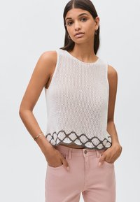 Bílý pletený svetr s žebrovanou texturou, bez rukávů, s vzorovaným hnědým lemem na dolním okraji, doplněný světle růžovými kalhotami.