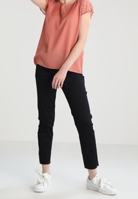 Jasnoróżowa bluzka z koronkowymi akcentami, zestawiona z czarnymi dopasowanymi spodniami i białymi trampkami. Bluzka ma okrągły dekolt i krótkie rękawy.