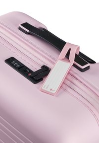 Valise rigide rose avec une surface texturée, une poignée de transport noire, un cadenas à combinaison et une étiquette de bagage rose amovible pour l'identification.
