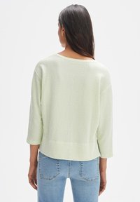 Hellgrüner Strickpullover mit strukturiertem Muster, weitem Ausschnitt und dreiviertel Ärmeln, kombiniert mit hellblauen Jeans.