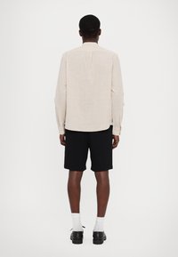 Camicetta beige chiaro a maniche lunghe con linee verticali, abbinata a pantaloni corti neri. La camicia ha un colletto a punte e polsini bottonati.